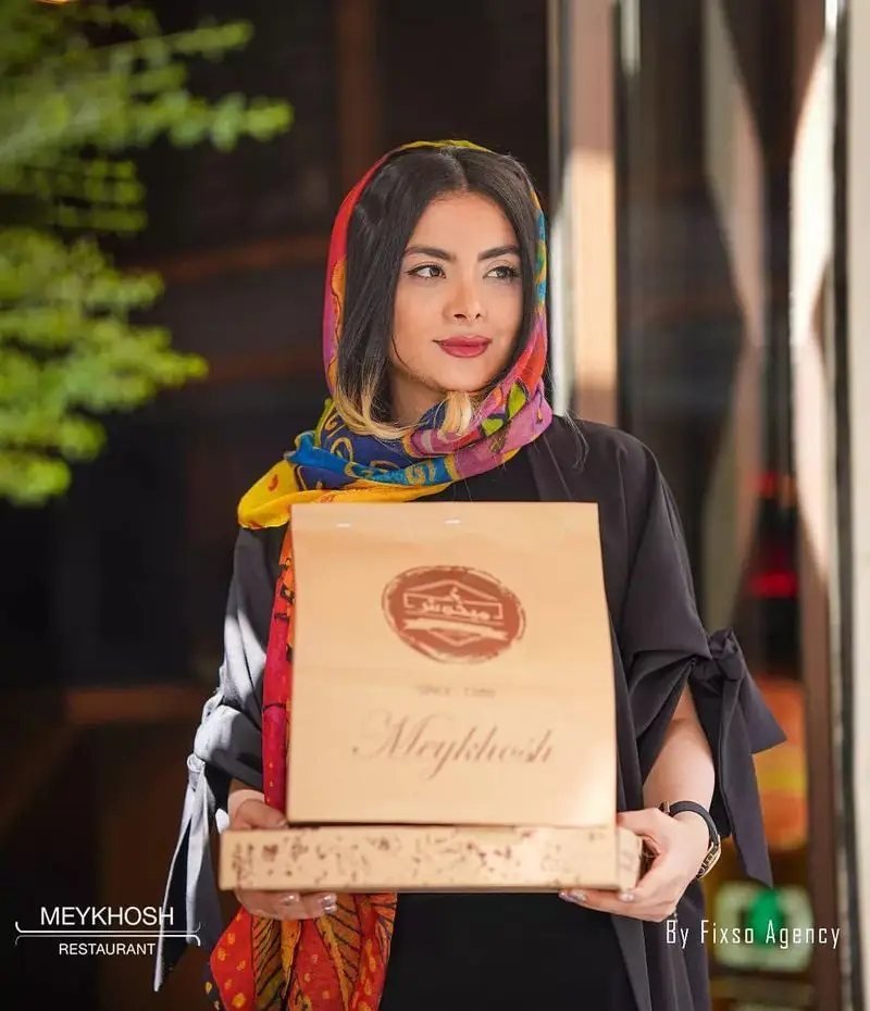 بانوی شیک‌پوش با روسری رنگارنگ، بسته‌بندی غذای فست فود میخوش را حمل می‌کند، نمادی از کیفیت و سرویس عالی رستوران میخوش.