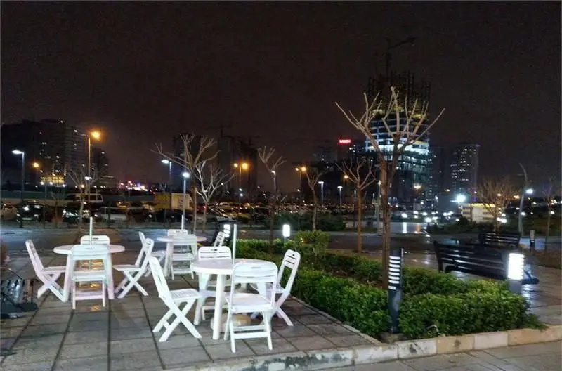 فضای باز دلنشین فست فود پرگوک در شب با میز و صندلی‌های سفید، درختان و نورپردازی جذاب شهری، ایده‌آل برای یک شام به‌یادماندنی.