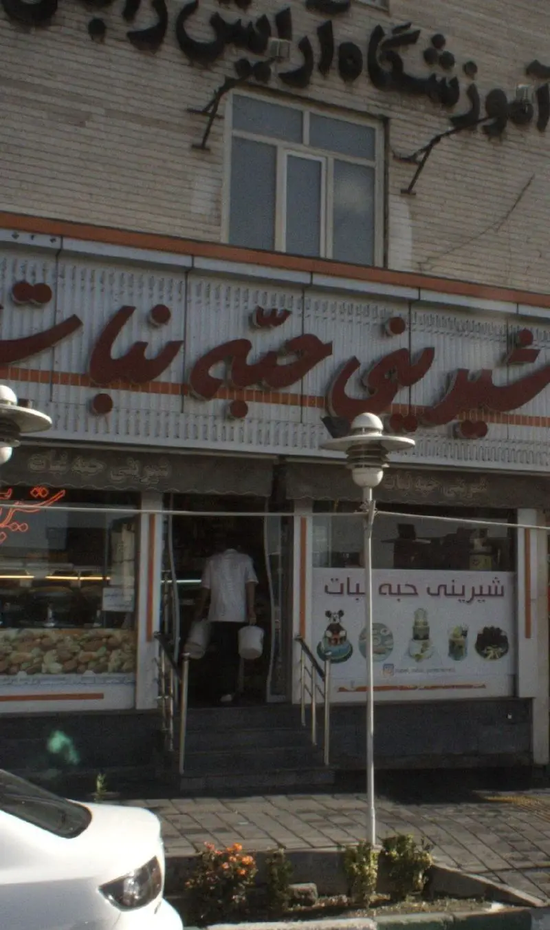 تصویر نمای یک شیرینی فروشی به نام شیرینی حبه نبات، با تابلو بزرگ و ویترین پر از انواع شیرینی و دسر خوشمزه.