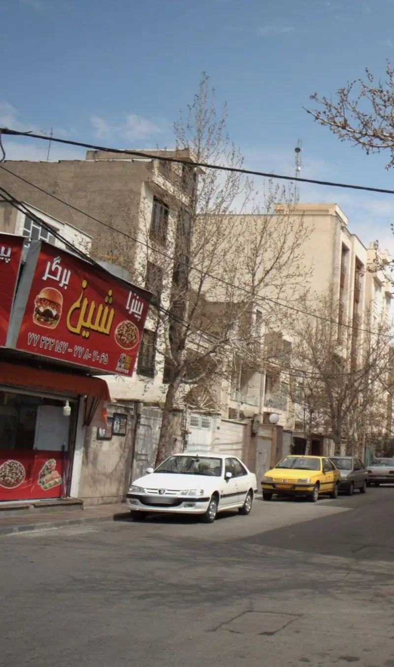 تصویر خیابانی در تهران با تابلوی پیتزا شین که انواع برگر و پیتزا ارائه می‌دهد، همراه با ساختمان‌ها و خودروهای پارک شده در حاشیه خیابان.