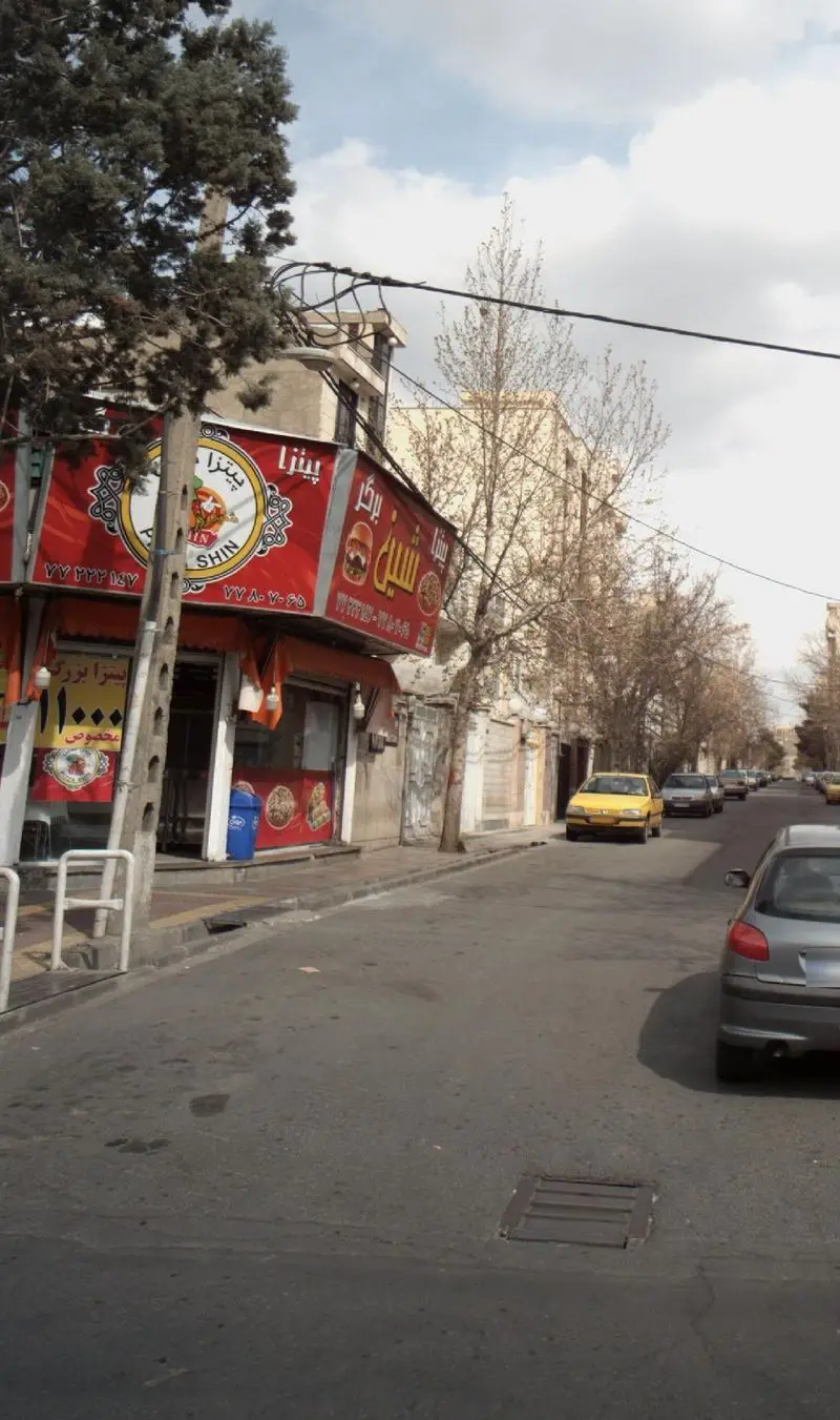 تصویری از نمای خیابان جلوی فست فود پیتزا شین و برگر در تهران، با تابلوی قرمز بزرگ، خودروهای پارک شده و درختان خیابانی.