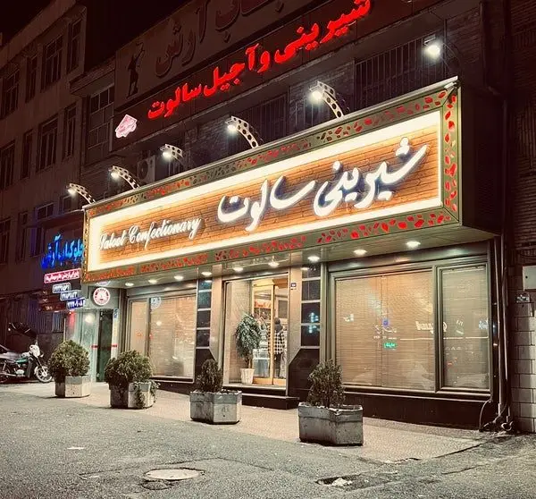 تصویری از نمای شب شیرینی و آجیل سالوت تهرانپارس، یک قنادی مدرن و زیبا با تابلوی روشن و ویترین‌های جذاب در مرکز شهر.