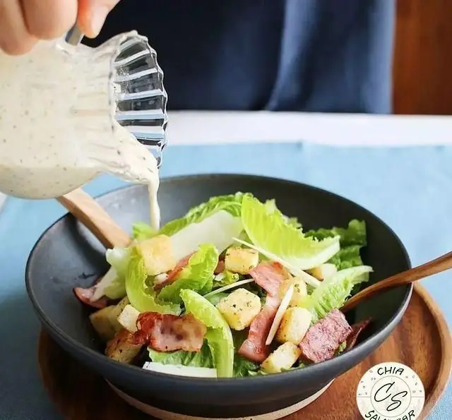 ریختن سس سفید خامه‌ای بر روی سالاد سزار خوشمزه با کاهو تازه، تکه‌های نان کروتون طلایی و بیکن ترد، در کاسه‌ای شیک از سالادبار چیا.