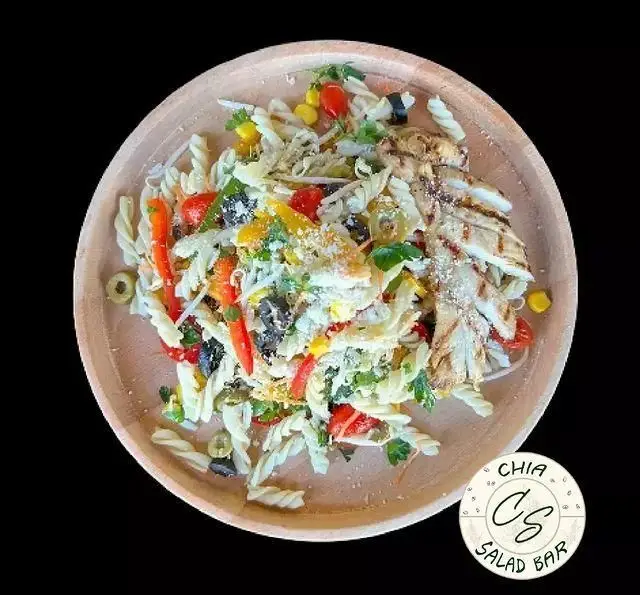 سالاد پاستا فوسیلی رژیمی با تکه‌های مرغ گریل شده، سبزیجات تازه، زیتون و ذرت، با چاشنی پنیر، از سالادبار چیا در بشقابی چوبی.