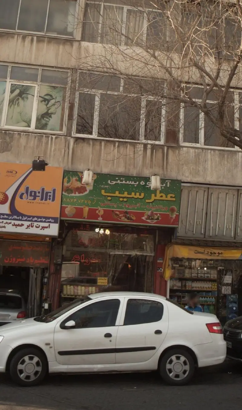 تصویر نمای خیابانی از مغازه آبمیوه و بستنی عطر سیب با تابلوی سبز روشن، یک خودروی سفید رنگ در جلو و درختان بدون برگ در بالای آن.