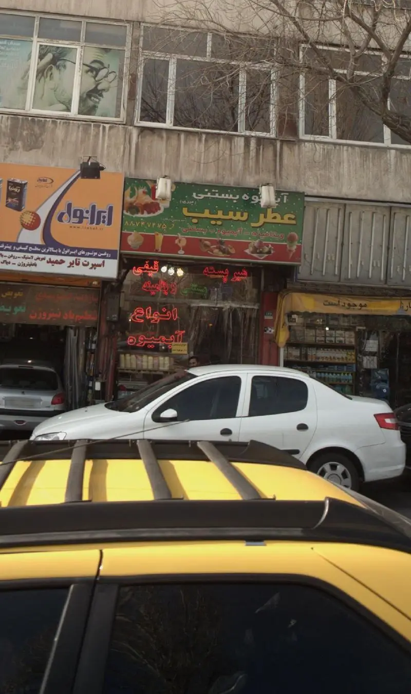 تصویر یک خیابان شهری با مغازه آبمیوه بستنی عطر سیب، تابلوهای تجاری از جمله ایرانول و ساختمان‌های بالا، همراه با خودروها.