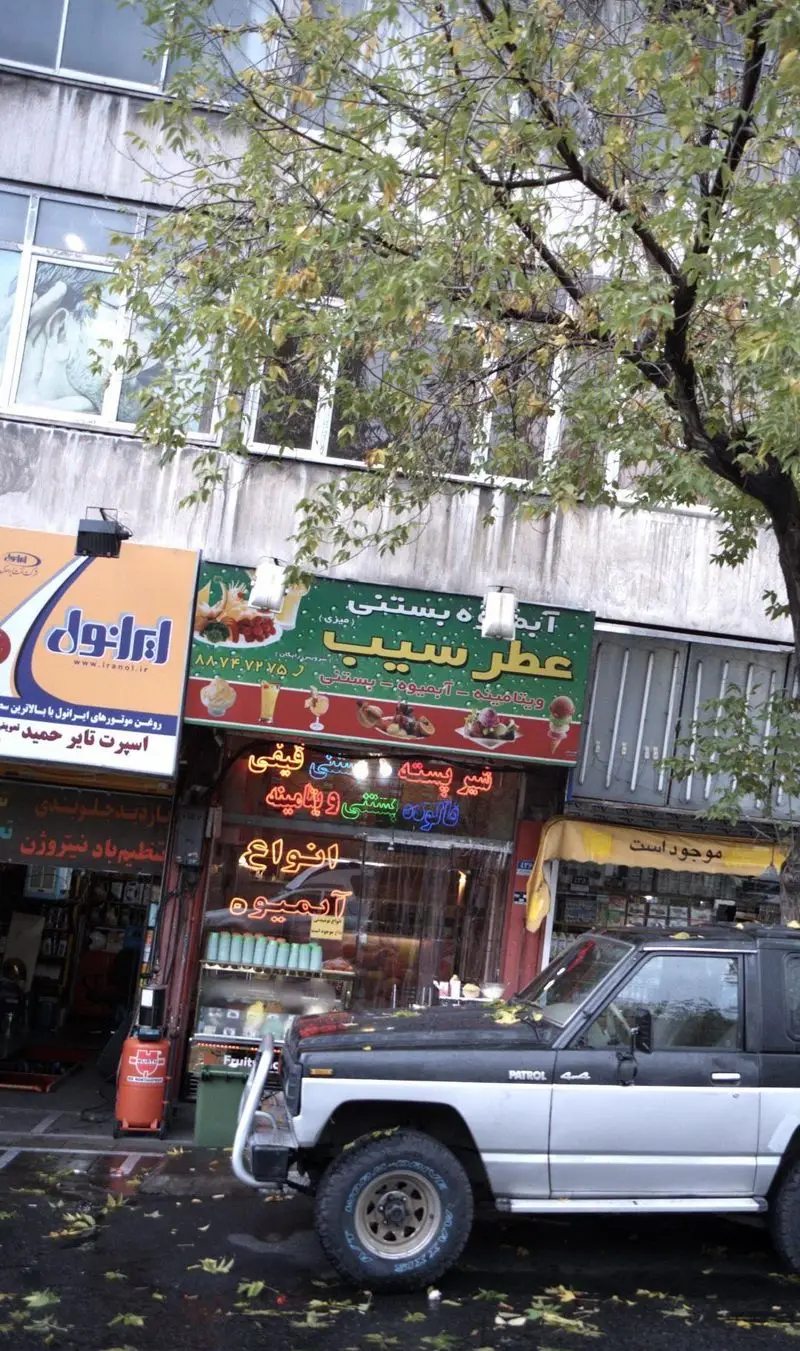 نمای خارجی مغازه آبمیوه بستنی عطر سیب در یک خیابان پاییزی، با درختان برگ‌ریز و خودروی پاترول پارک شده در جلو.