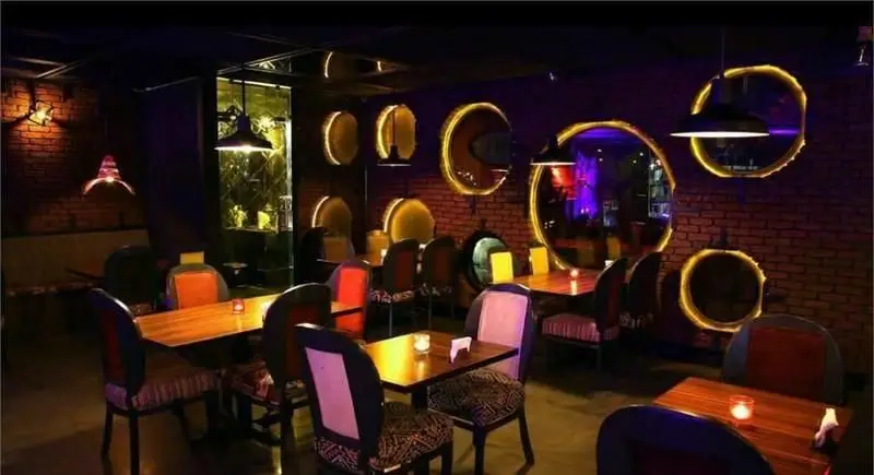 تصویری از فضای دلنشین کافی شاپ جیلیز، با دکوراسیون مدرن، نورپردازی خاص و آینه‌های نورانی دایره‌ای روی دیوارهای آجری