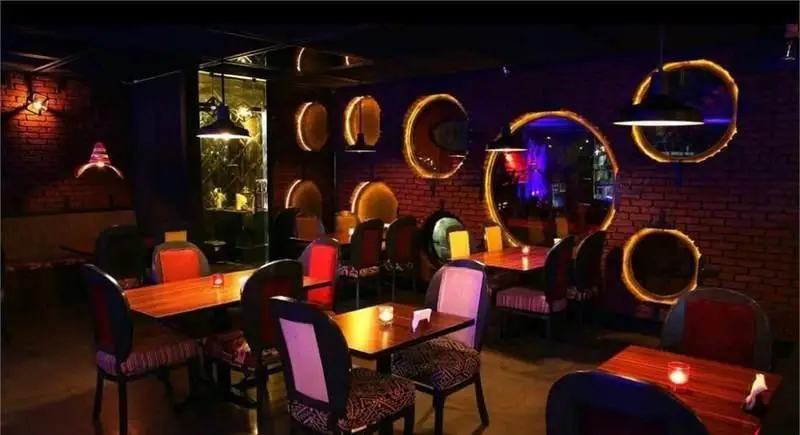فضای داخلی کافی شاپ جیلیز با دکوراسیون مدرن، نورپردازی خاص، میز و صندلی های شیک و آینه های گرد روشن بر روی دیوار آجری.
