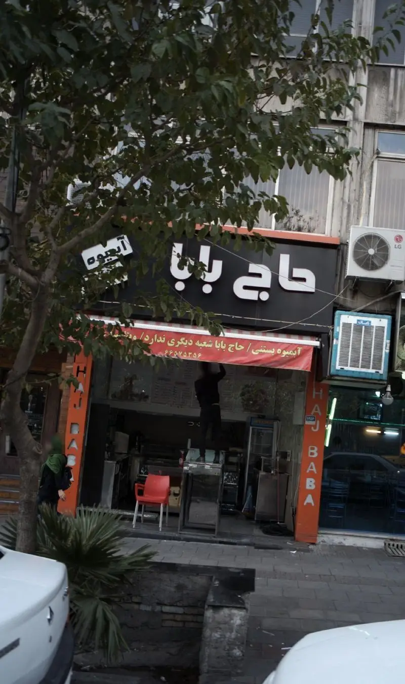 آبمیوه بستنی حاج بابا، فروشگاهی پر جنب و جوش در خیابان. تابلوی بزرگ و سفید، با درخت سبز و تردد شهری.