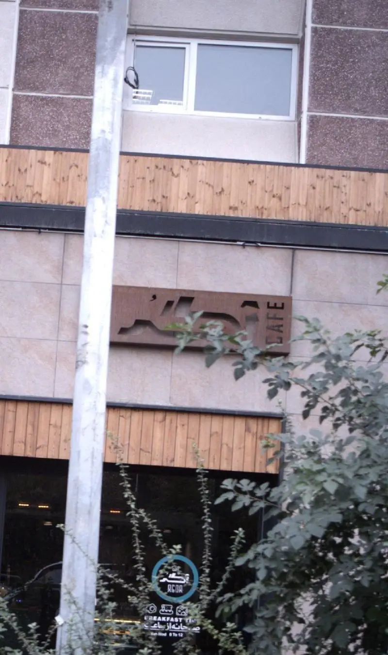 نمای بیرونی کافه آگر، با تابلوی قهوه‌ای Agor Cafe و جزئیات زمان صبحانه ۸ تا ۱۲ روی شیشه. طراحی مدرن ساختمان با نمای چوبی و کاشی.