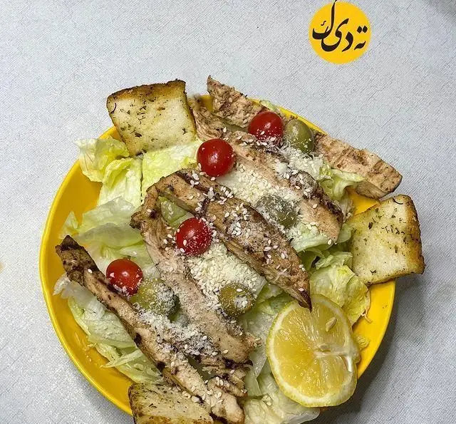 سالاد سزار خوشمزه با مرغ گریل آبدار، کاهو تازه، گوجه گیلاسی، زیتون، نان کروتون و لیمو در رستوران ته دیگ بام لند