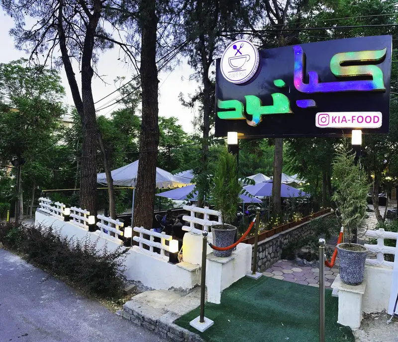 نمایی از ورودی باغ رستوران و کافه کیافود (کافه کیا) با تابلوی روشن، نورپردازی شبانه، فضای سبز و آلاچیق‌های جذاب.