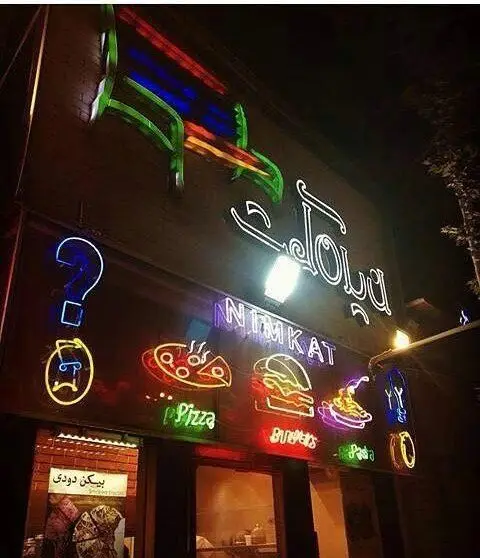 تصویری جذاب از نمای بیرونی رستوران ایتالیایی نیمکت در شب، با تابلوهای نئونی پیتزا، برگر، پاستا و بیکن دودی.