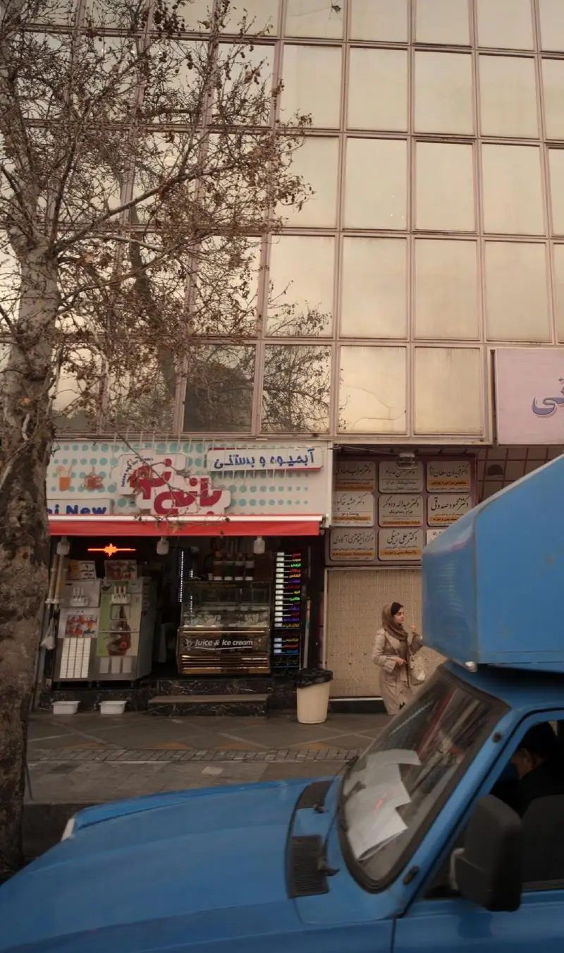 تصویری از فروشگاه آبمیوه و بستنی بانی نو با ویترین رنگارنگ، درختی در کنار و ماشینی آبی در حال عبور در خیابان.