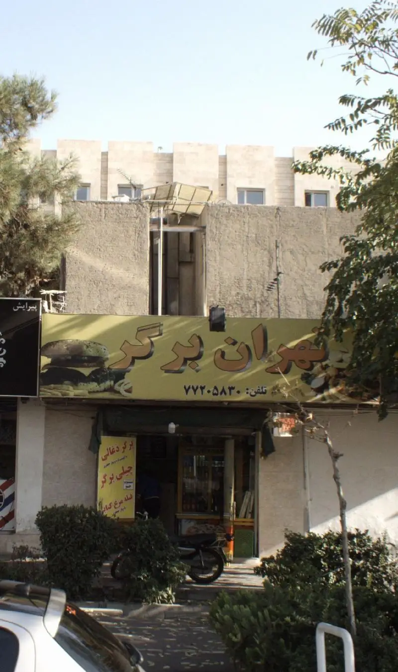 نمای بیرونی فست فود تهران برگر، تابلوی زرد با تصویر همبرگر، شماره تماس ۷۷۲-۰۸۳۰ و منوی برگر ذغالی و چیلی برگر.