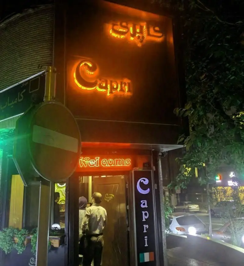 ورودی رستوران ایتالیایی کاپری در شب، با تابلوهای نئونی "Capri" و "کاپری" و "Welcome". پرچم ایتالیا و مشتریان در حال ورود.
