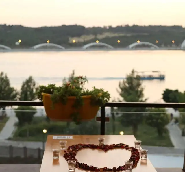 تصویری زیبا از کافه رستوران رولینجا: میز چیدمان رمانتیک با قلب گل رز و لیوان شات، مشرف به منظره آب و پل در افق.