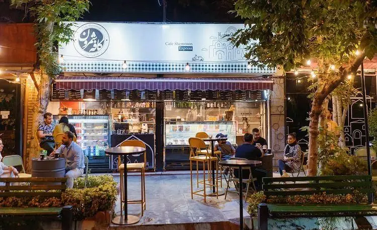 نمای بیرونی کافه شمرون اکسپرس در شب با مشتریان در فضای باز و نورپردازی دلنشین. پاتوقی دنج برای دورهمی و لذت بردن از نوشیدنی.