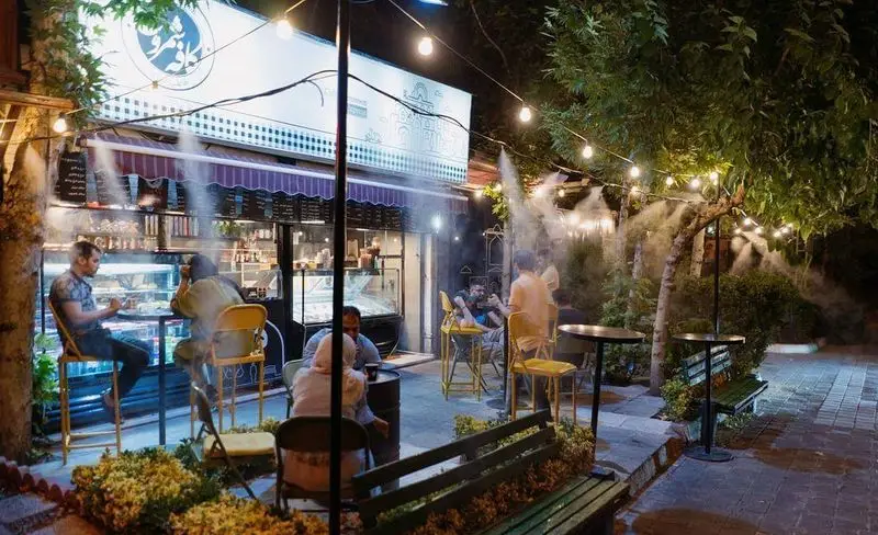 نمایی شبانه از کافه رستوران شمرون با فضای باز دلنشین، میز و صندلی، سیستم مه پاش خنک کننده و ریسه‌های نورانی. مشتریان در حال لذت بردن از محیط.