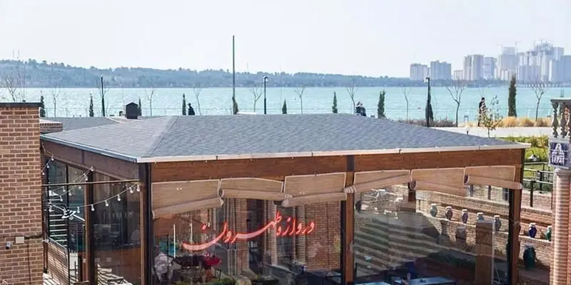 رستوران ایرانی دروازه طهرون با نمای مدرن چوبی و شیشه‌ای و سقف شیب‌دار، واقع در کنار دریاچه با چشم‌انداز زیبای شهری تهران.