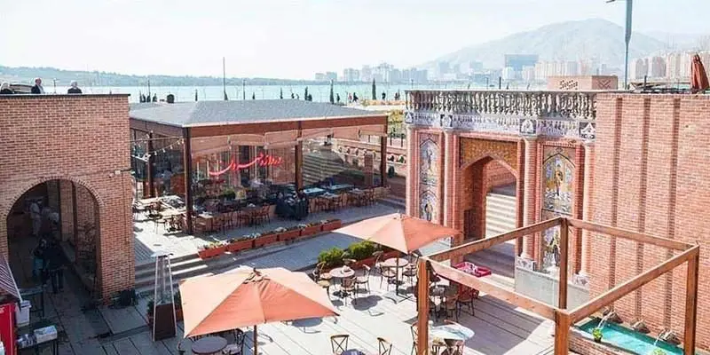 رستوران ایرانی دروازه طهرون با فضای باز دلباز، بنای سنتی و شیشه‌ای مدرن، محوطه غذاخوری روباز و چشم‌انداز زیبای دریاچه و کوهستان.