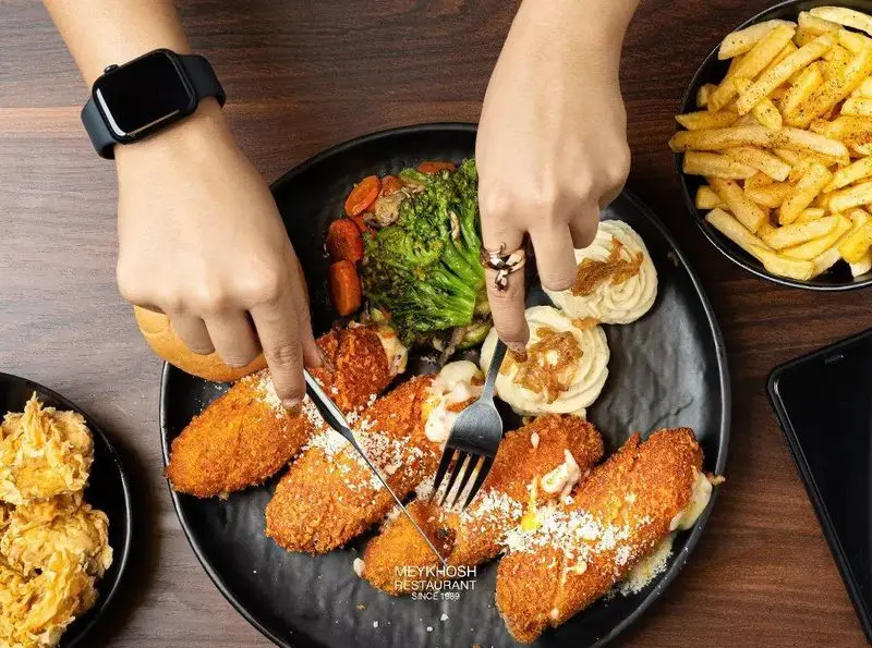 نمایی از دست‌ها در حال برش کوردن بلو با پنیر ذوب شده، سبزیجات تازه، پوره سیب‌زمینی و سیب‌زمینی سرخ‌کرده. فست فود میخوش.