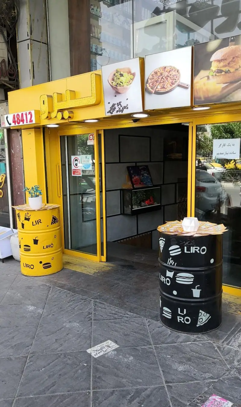نمای ورودی فست فود لیرو با رنگ زرد، تابلوهای غذای آماده شامل پیتزا، برگر، سالاد و میزهای بشکه ای خلاقانه.