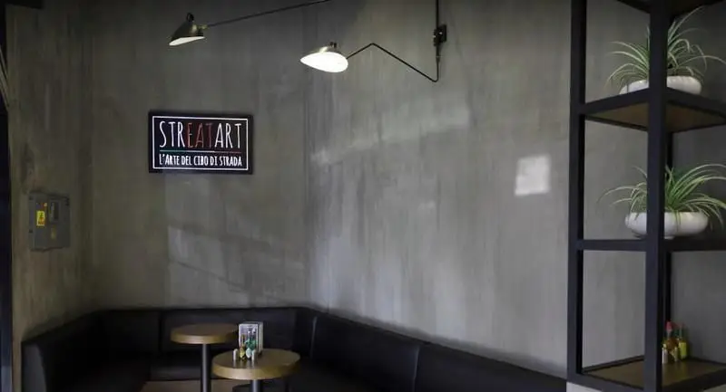 فضای داخلی مدرن و شیک پیتزایی استریت آرت با دیوار بتنی، میز و نیمکت‌های سیاه، نورپردازی هنری و قفسه‌های تزئینی گیاه.