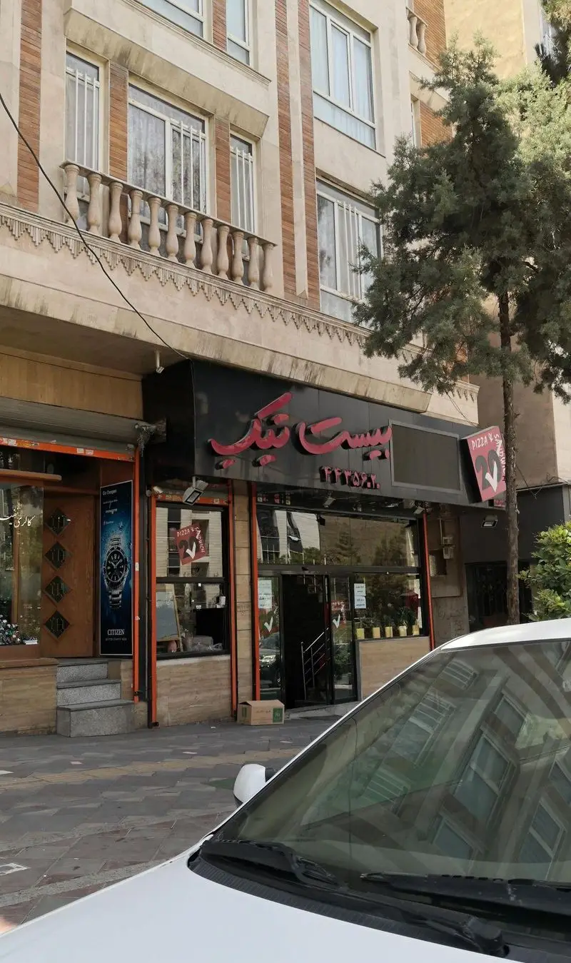 نمای بیرونی فست فود بیست تیک با تابلوی بزرگ قرمز، پیتزا و ساندویچ فروشی در خیابان، با ساختمان مسکونی در پس‌زمینه و خودرویی در پیش‌زمینه.