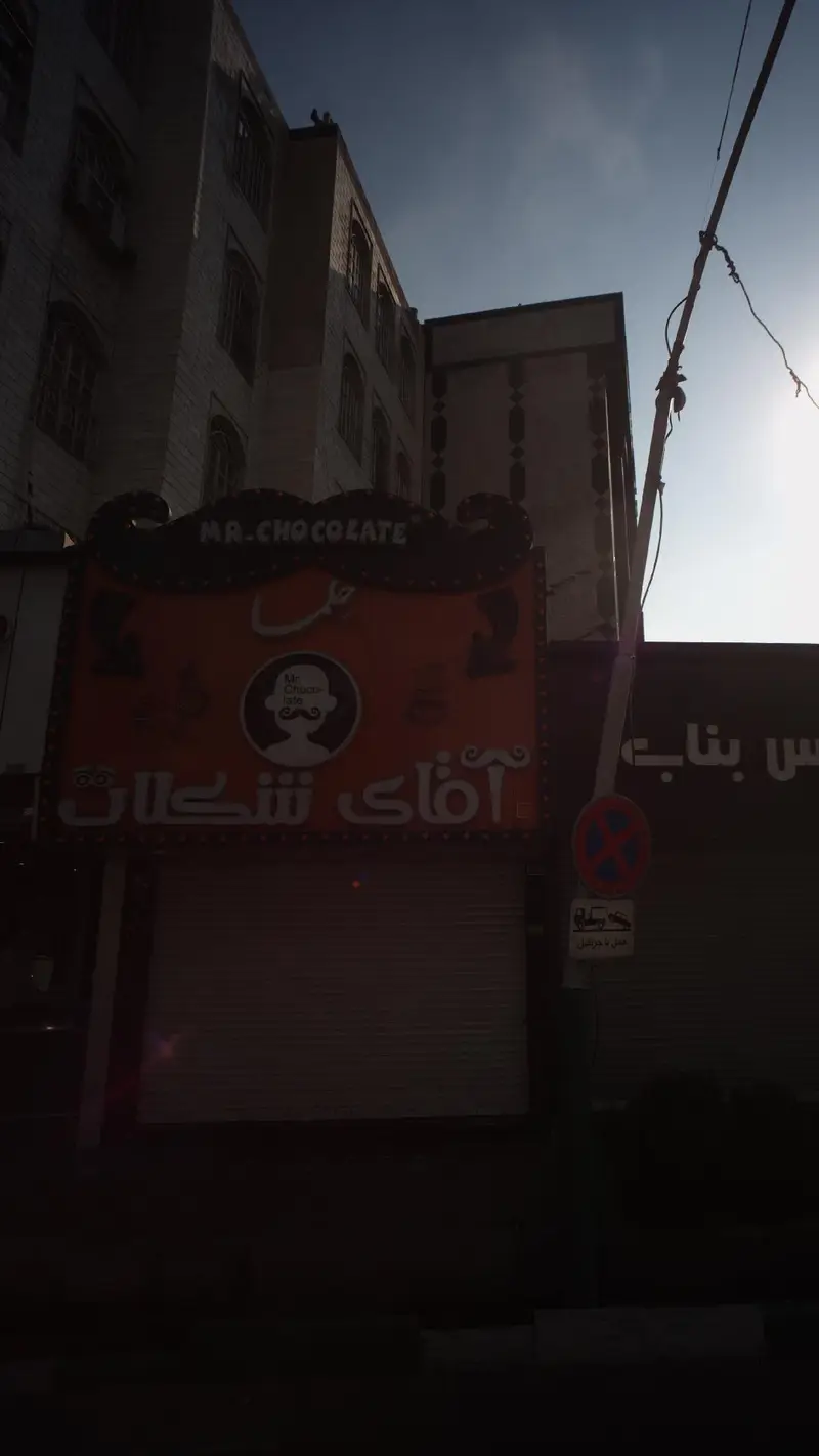 نمای بیرونی فروشگاه حلما آقای شکلات با تابلوی بزرگ «Mr. Chocolate» و لوگوی مرد سبیل‌دار، کرکره‌های فروشگاه پایین است.