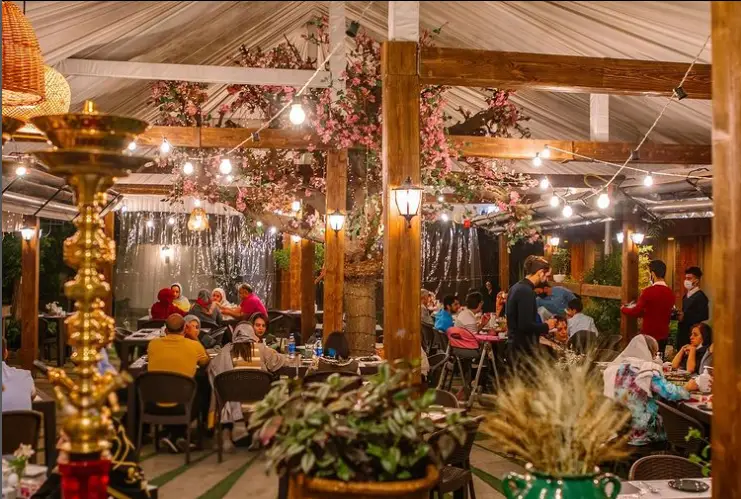 فضای دلنشین رستوران گیلانه با مشتریانی در حال صرف غذا، دکوراسیون چوبی، نورپردازی گرم و شکوفه‌های صورتی، قلیان در پیش‌زمینه.