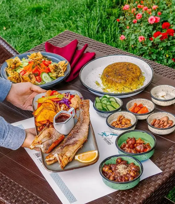غذای خوشمزه شمالی رستوران گیلانه: ماهی سرخ شده، باقالی پلو، سالاد و مخلفات گیلانی نظیر زیتون پرورده و سیر ترشی در فضای سبز