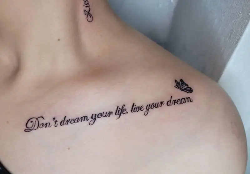 تصویری از تاتوی متنی روی ترقوه و شانه یک فرد با جمله "Don't dream your life, live your dream" و طرح پروانه کوچک. این تاتو با فونت زیبا و ظریف اجرا شده است.