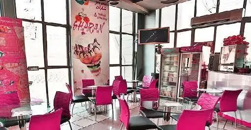 تصویری از فضای داخلی فست فود شاران با دکوراسیون مدرن، صندلی‌های صورتی، میزهای شیشه‌ای و تابلو بزرگ منوی جذاب.