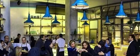 محیط دنج و پر جنب و جوش کافی شاپ تومو با مشتریان متعدد و دکوراسیون مدرن شامل چراغهای آویز آبی و قفسههای شیک.
