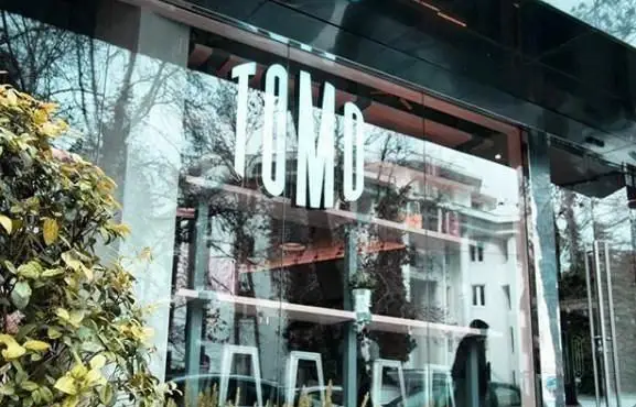نمایی جذاب از کافی شاپ تومو با دکوراسیون شیشه ای مدرن، نام "TOMO" به وضوح روی شیشه و انعکاس زیبای فضای سبز و شهری.