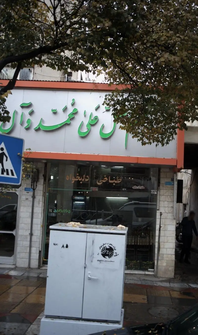 تصویر نمای بیرونی طباخی دلبخواه با سردر آش علی محمد ذوال، یک غذاخوری محلی در خیابانی با درختان پاییزی و علائم شهری.