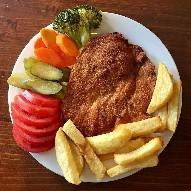 بشقاب شنیتسل لذیذ کافه لینت شامل کتلت مرغ برشته، سیب زمینی سرخ کرده و سبزیجات تازه مانند گوجه، خیارشور و بروکلی.