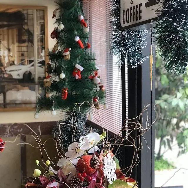 دکوراسیون کریسمس کافه لینت: درخت سبز با جوراب و کلاه کوچک، گل‌آرایی با ارکیده و میوه کاج، تابلوی قهوه، فضای دنج برای سال نو.