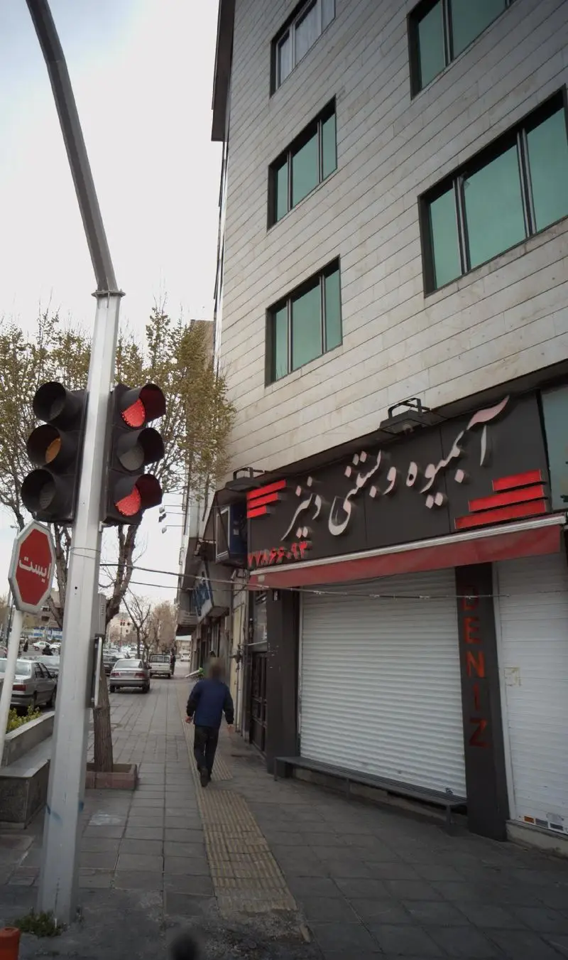 مغازه آبمیوه و بستنی دنیز با کرکره‌های پایین در خیابان، همراه با چراغ راهنمایی قرمز، تابلو ایست، ساختمان بلند و عابر پیاده در پیاده‌رو شلوغ.