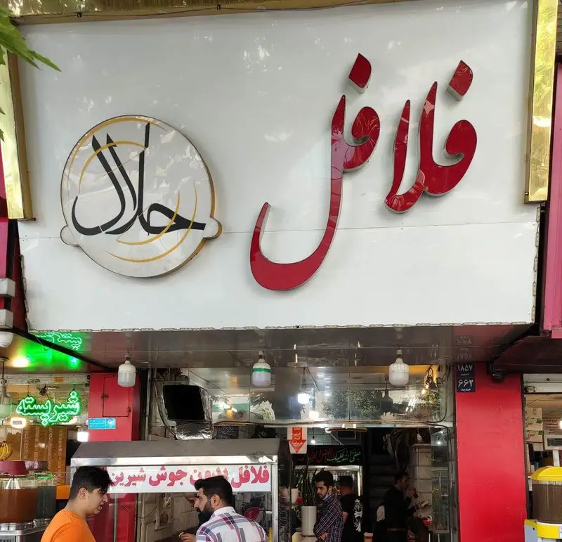 تصویری از نمای بیرونی مغازه فلافل حلال با تابلوی بزرگ و مشتریان در حال خرید، نشان‌دهنده یک کسب و کار غذایی اسلامی.