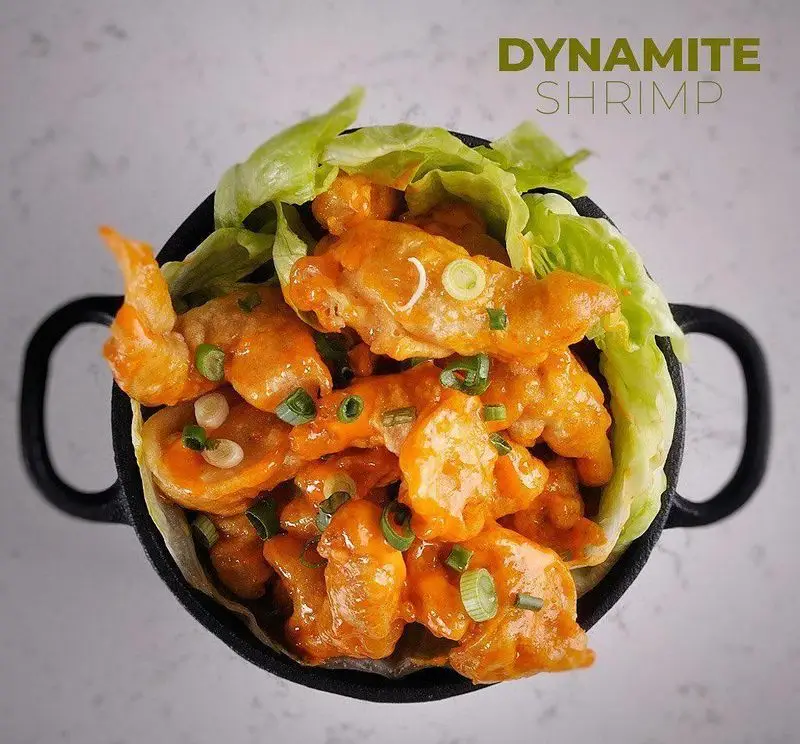 داینامیت شرمپ خوش‌رنگ و لذیذ رستوران بامبو اکسپرس؛ میگوهای سوخاری با سس نارنجی، پیازچه و کاهو در کاسه مشکی. غذای آسیایی پرطرفدار.