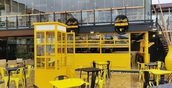 نمایی جذاب از فست فود کیوسک برگر با طراحی مدرن و رنگ‌های شاد زرد و مشکی، همراه با فضای باز دلپذیر و میز و صندلی.