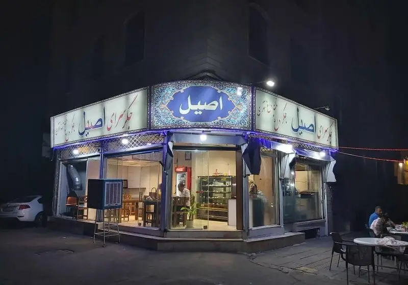 نمای شبانه روشن از جگرسرای اصیل، رستورانی محبوب برای جگر و کباب، با تابلوی آبیرنگ، میزهای فضای باز و داخل مغازه با کارکنان.