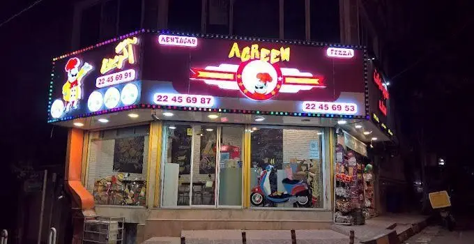 نمای بیرونی فست فود آگرین در شب، با تابلوی نئونی درخشان که نام آگرین، کلمات کنتاکی و پیتزا را با لوگوی سرآشپز نمایش می‌دهد. شماره تماس‌ها نیز قابل مشاهده است.