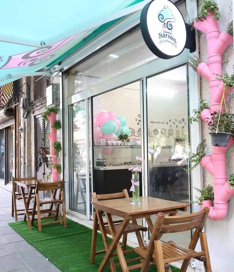 کافه قنادی نارون با نمای بیرونی جذاب، میزهای چوبی شیک، چمن مصنوعی، دکوراسیون صورتی و سایه‌بان فیروزه‌ای. محیطی دلنشین برای مشتریان.