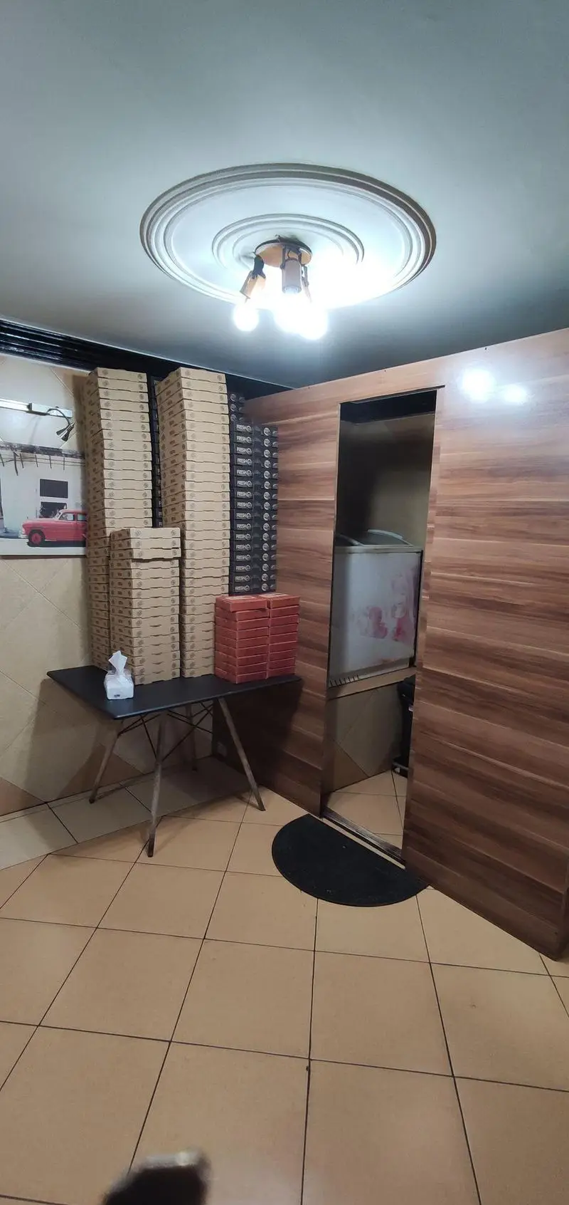 فضای داخلی فست فود جزیره با انبوهی از جعبه‌های پیتزای آماده، یک میز سیاه، و یخچالی در کنار دیوار چوبی. آماده برای ارائه بهترین پیتزاها.