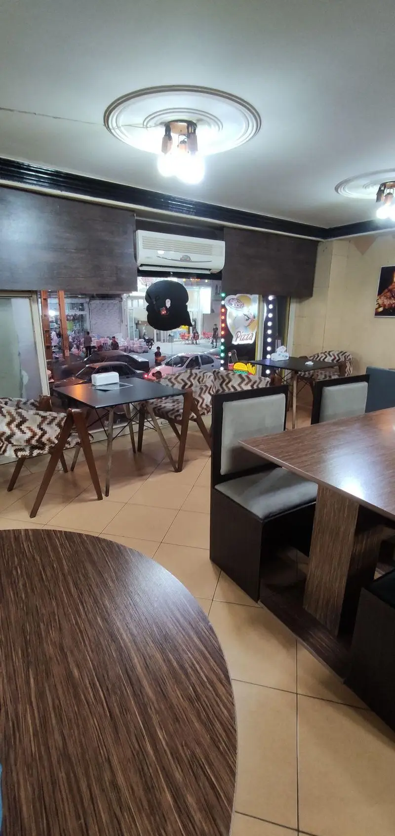 فضای داخلی دنج و مدرن فست فود جزیره با میز و صندلی‌های چوبی، پنجره رو به شهر و نوری دلپذیر، محیطی عالی برای تجربه غذایی لذت‌بخش.