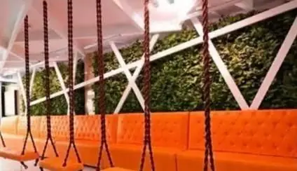 تصویری از طراحی داخلی منحصر به فرد کافی شاپ سیمو شامل نشیمن‌های نارنجی، تاب‌های معلق و دیوار سبز مدرن با الگوهای هندسی سفید برای فضایی آرام و شیک.